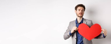 Tutkulu yakışıklı adam kırmızı sevgililer günü kartlarıyla kalp atışları yapıyor. Takım elbise içinde duruyor ve aşkı arıyor. Beyaz arka planda duruyor..