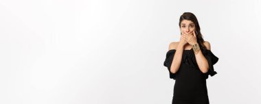 Moda ve güzellik. Siyah elbiseli çekici bir kadın ağzı kapalı ve nefes nefese, endişeli kameraya bakıyor, beyaz arka planda dikiliyor..