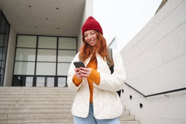Redhead girl, young woman tourist with backpack, holds smartphone, looks for route on mobile application, searches for hotel on phone map, smiles happily.