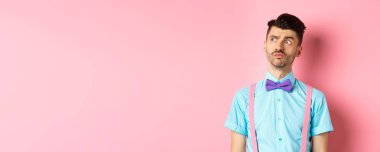 Romantik kıyafetli dalgın genç adam, başka tarafa bakıyor ve düşünüyor, pembe arka planda dikiliyor süslü papyonlu ve gömlekli.