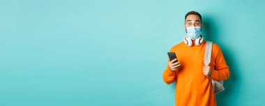 Yüz maskeli genç adam cep telefonu kullanıyor, sırt çantası taşıyor, kameraya bakıyor, açık mavi arkaplana karşı duruyor..
