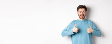 Heyecanlı gülümseyen adam mükemmel ürünü övmek için başparmağını kaldırıyor, kameraya şaşkın ve mutlu bakıyor, iyi bir anlaşma öneriyor, beyaz arka planda duruyor..