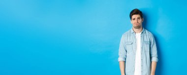 Kameraya şüpheyle bakan, somurtan, somurtan, bir şey hakkında huzursuz hisseden, mavi arka plana karşı duran bir adam..