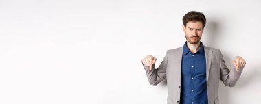 Hüsrana uğramış sakallı erkek girişimci olumsuz duygularla somurtuyor ve işaret ediyor, beyaz arka planda duran hoşnutsuzluğunu ya da kötü düşüncelerini dile getiriyor..