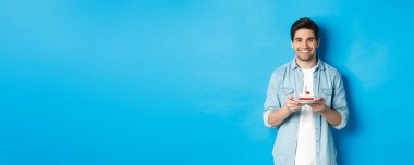 Gülümseyen adam doğum gününü kutluyor, elinde mumla doğum günü pastası tutuyor, mavi arka planda duruyor..