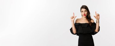 Moda ve güzellik. Siyah elbiseli düşünceli, çekici kadın dik dik bakıyor ve işaret ediyor, baştan çıkarıcı bakışlarla düşünüyor, beyaz arka planda..