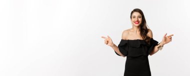 Moda ve güzellik. Mücevher, makyaj ve siyah elbise giymiş çekici bir kadın, gülümseyen ve parmaklarını yana doğru gösteren fotokopi alanı teklifi, beyaz arka plan.
