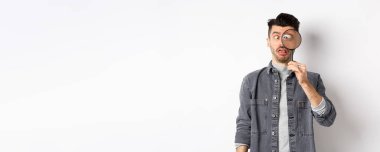 Komik genç adam gözlerini kısıyor, büyüteçle yüzlerini gösteriyor, beyaz arka planda duruyor..