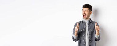 Komik yakışıklı adam göz kırpıyor ve kamerayı işaret ediyor, seni tanıtıma davet ediyor, beyaz arka planda duruyor..