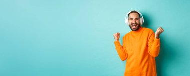 Turuncu süveter giyen mutlu yetişkin adam, yukarı bakıyor ve kulaklıkla müzik dinliyor, mavi arka planda duruyor..