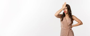 Cazibeli, şık bir kadın facepalm yapıyor, gözlerini yuvarlıyor, aptalca bir şeyden rahatsız oluyor, beyaz arka planda bir partide duruyor..