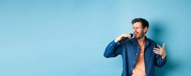 Romantik adam mikrofon ve jest ile şarkı söylüyor, karaoke yapan şarkıcı, mavi arka planda duruyor..