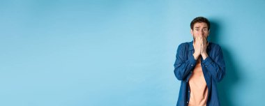 Korkmuş endişeli adam elleriyle ağzını kapatıyor ve mavi arka planda duran korkunç bir şeye bakıyor..