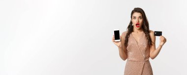 İnternetten alışveriş ve tatil konsepti. Kadın şaşırmış görünüyor ve beyaz arka planda duran akıllı telefon ekranlı kredi kartı gösteriyor..