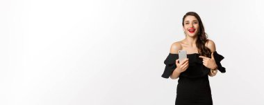 İnternetten alışveriş konsepti. Siyah elbiseli tatmin olmuş, gülümseyen, mutlu ve beyaz arka planda duran, cep telefonunu işaret eden güzel bir kadın..