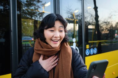 Portrait of cheerful asian girl talks on mobile phone, video chats, looks amazed at smartphone camera, stands on bus stop.