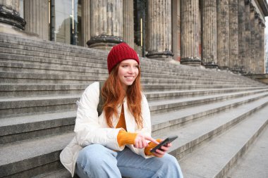 Stylish young redhead woman, talking on mobile phone app, using social media application, looking for something online on smartphone, sits on stairs outdoors.