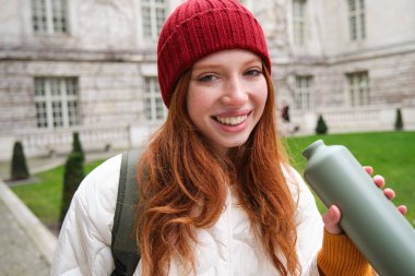 Portrait of happy girl traveler, drinking hot tea from thermos while walking and exploring city. Tourist with flask smiling at camera.