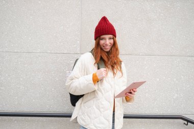 Young redhead woman with red hat, uses her digital tablet outdoors, stands on street with gadget, connects to wifi internet and searches for a location in internet.