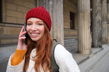 Smiling redhead female tourist talks on mobile phone and walks around city. Happy student in red hat calls friend, stands on street and uses smartphone.