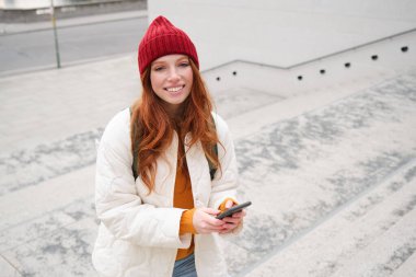 Smiling redhead girl, student tourist walks around city, goes up the stairs, looks at mobile phone map to get around town, sends message on smartphone.
