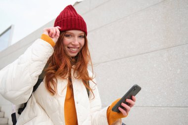 Joyful girl tourist looks at mobile phone, texts message on smartphone social media application, walks around city, looks for sightseeing on mobile app.