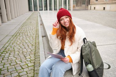 Redhead girl smiles, sits outdoors near building with digital tablet, thermos and backpack, connects to public internet and searches smth online on her gadget.