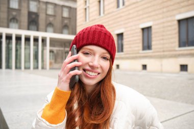 Mobile connection and people concept. Happy redhead woman in hat, talks on mobile phone, making telephone call, using app to call abroad.