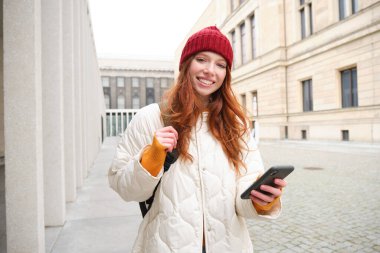 Beautiful smiling girl, tourist with backpack, holding smartphone, using map on mobile phone application, looking for sightseeing in internet app, standing outdoors.