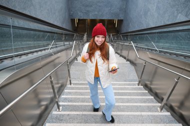 Outdoor shot of young woman plans route, follows map on smartphone app, goes up stairs with backpack and smiles.