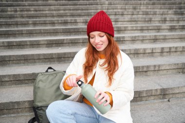 Smiling tourist, girl sits on stairs, rests on staircase, takes thermos from backpack, drinks hot coffee from flask.
