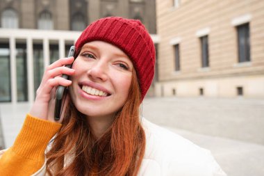 Mobile connection and people concept. Happy redhead woman in hat, talks on mobile phone, making telephone call, using app to call abroad.