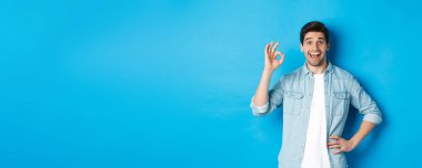 Gülümseyen mutlu adam işaret veriyor ve memnun görünüyor, iyi bir şeyi onaylıyor, mavi arka plana karşı duruyor..