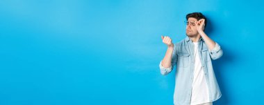 Gözlüklü genç adam tırnaklarına bakıyor, maniküre bakıyor, mavi arka planda duruyor..