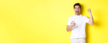 Akıllı telefondan kazanan neşeli adam, elini kaldıran ve cep telefonunu tutan, uygulama hedefine ulaşan, sarı arka planda duran.