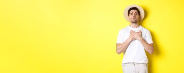 Turizm ve yaz kavramı. Hasır şapkalı romantik adam nostaljik görünüyor, gözleri kapalı ve kalbi üzerinde el ele tutuşuyor, sarı arka planda duruyor..