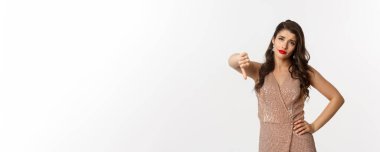 Şık elbiseli, kuşkucu genç kadın hayal kırıklığına uğramış görünüyor, baş parmağını kaldırıyor, Noel partisinden hoşlanmıyor, beyaz arka planda duruyor..