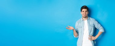 Dikkatsiz bir adam hayal kırıklığı yaratan bir şeye bakıyor, şikayet ediyor ve kötü ürünü yargılıyor, mavi arka planda eğlenmeden duruyor..