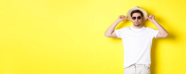 Tatile hazır, hasır şapka ve güneş gözlüğü takmış, sarı arka planda duran kendinden emin genç turist..