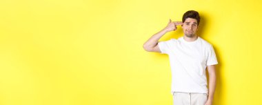 Sıkıntılı adam kendini parmak tabancasıyla vuruyor, hoşnutsuz ve yorgun görünüyor, sarı arka planda duruyor..