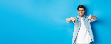 Hayal kırıklığına uğramış bir adam, kötü bir ürüne, kötü reklama, mavi arka plana karşı duran, kötü bir ürüne burun kıvırıyor ve yüzünü buruşturuyor..