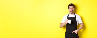 Siyah önlüklü, gülümseyen genç barista elinde kahve fincanı, sarı arka planda duruyor..
