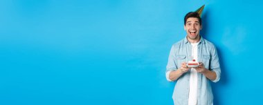 Neşeli genç adam doğum gününü parti şapkasıyla kutluyor, doğum günü pastası tutuyor, mavi arka planda duruyor..