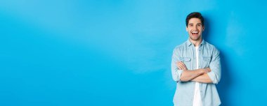 Sakallı, sıradan giyinmiş, komik bir şeye gülüp bakan, mavi arka planda duran yakışıklı bir adam..