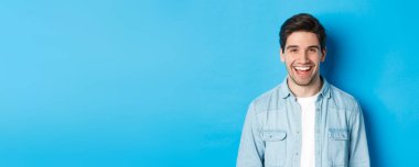 Yakışıklı genç adam gülüyor, günlük kıyafetler giyiyor, mavi arka planda dikiliyor..