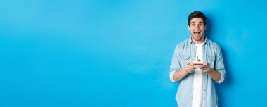 Doğum günü pastası tutan ve kameraya gülümseyen heyecanlı adam mavi arka planda doğum gününü kutluyor..