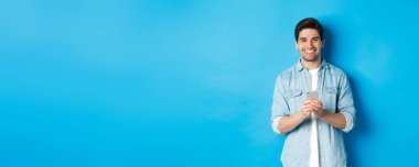 Sıradan kıyafetli yakışıklı adam kameraya gülümsüyor, akıllı telefonu kontrol ediyor, mavi arka planda duruyor..