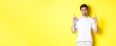 Komik olmayan yakışıklı adam kaşlarını çatıyor, kötü bir şeyi işaret ediyor, sarı arka plana şüpheyle bakıyor..