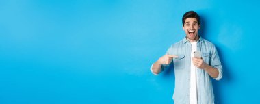 Çevrimiçi alışveriş, uygulama ve teknoloji kavramı. Mutlu gülümseyen adam telefonu işaret ediyor, mavi arka plana hayret içinde bakıyor..