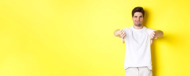 Şüpheci adam baş parmağını aşağı gösteriyor ve sırıtıyor, hoşlanmıyor ve katılmıyor, sarı arka planda duruyor. Boşluğu kopyala